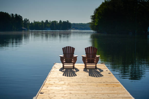 Adirondack chairs sitting on a wooden pier Two Adirondack chairs sitting on a wooden pier facing the calm water of a lake in Muskoka, Ontario Canada. A cottage nestled between trees is visible in background. promenade stock pictures, royalty-free photos & images