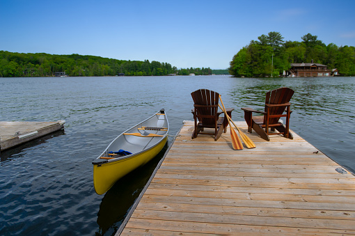 Two Adirondack chairs on a wooden pier facing the blue water of a lake in Muskoka, Ontario Canada. A yellow canoe is tied to the dock while the paddles are next to the chairs.