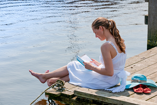high angle side view on a smiling woman with bare feet wearing a white sundress reading a book sitting on wooden pier at the waterfront