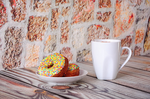 A plate with two fresh donuts in multi-colored sprinkles and a tall cup of cappuccino with ground cinnamon. Close-up.