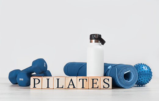 PILATES text. Rolled blue yoga mat and exercise resistance band dumbbells on grey wooden surface. Gender neutral fitness and exercise concept with copy space. Active lifestyle. Workout at home or gym