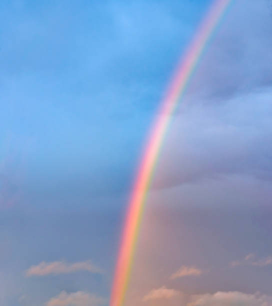 A part of a beautiful rainbow glows high up in the blue sky. Vibrant rainbow colours radiate and fluffy clouds float through the air. A full frame image.