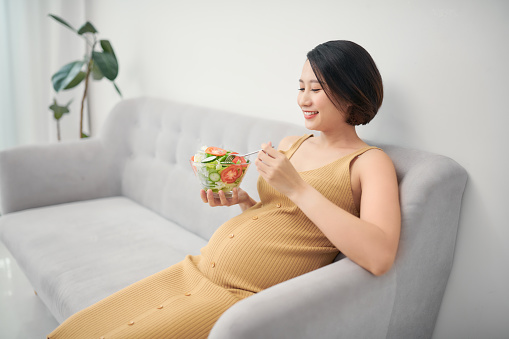 Pregnancy, healthy food and people concept. Happy pregnant woman eating vegetable salad for breakfast in sofa at home