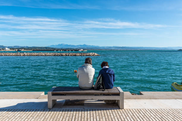 Katakolo, Greece - November 11, 2019: Couple rest on a seat on the waterfront, with the harbour in the background in Katakolo (Olimpia), Greece.