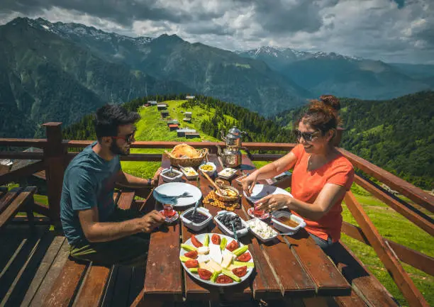 A young tourist couple eating delicious Turkish breakfast with various colorful foods and Black Sea Region food stretch muhlama or kuymak (Turkish Cheese Fondue) with nature landscape view at Pokut Highland in Çamlıhemşin, Rize in Karadeniz region of Türkiye