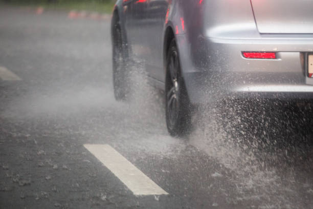 gli spruzzi d'acqua piovana scorrono da ruote di auto d'argento che si muovono velocemente nella città diurna con messa a fuoco selettiva. - sbandare foto e immagini stock