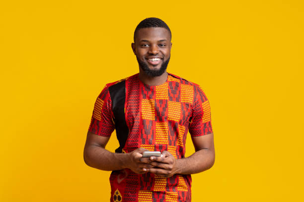 uśmiechnięty czarny facet za pomocą smartfona, ubrany w afrykański t-shirt - traditional style zdjęcia i obrazy z banku zdjęć