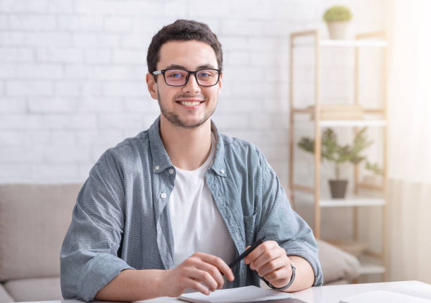 istock Online home study for students. Smiling guy with glasses sits at table in living room interior 1256457508