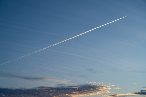 An airplane leaves a white vapor line on a beautiful blue sunset sky. Blue sky, dark clouds, and orange sunset