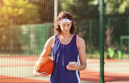 Serious young basketball player using smartphone at outdoor court after game