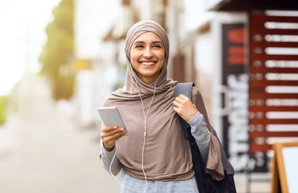 Cheery arab girl walking by city and listening to music, using smartphone, free space
