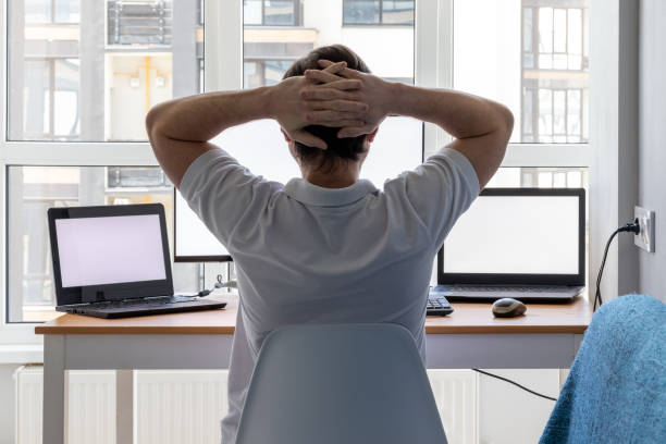 istock A man in a white t-shirt with hands folded behind his head is sitting at a workplace with two laptops and a monitor near the window. Remote work from home office. Back view 1256466568