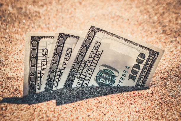istock Money dolars half covered with sand lie on beach close-up. 1256469224