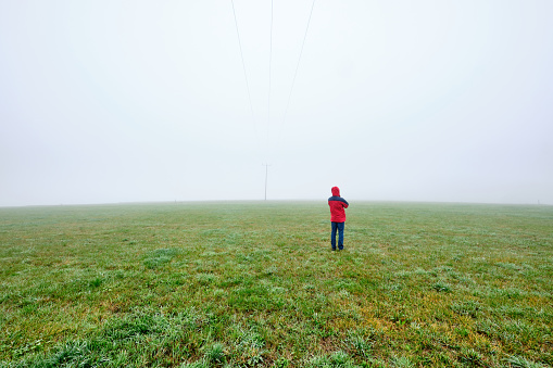 Rear view of man in red jacket standing on a green meadow and looking into a foggy nowhere landscape. Seen in October in Germany, Bavaria