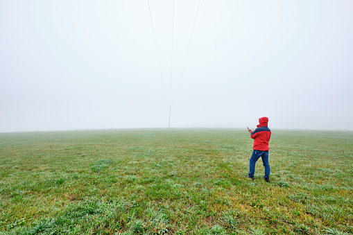 Rear view of man in red jacket standing on a green meadow and looking on his smartphone in front of a foggy nowhere landscape. Seen in October in Germany, Bavaria