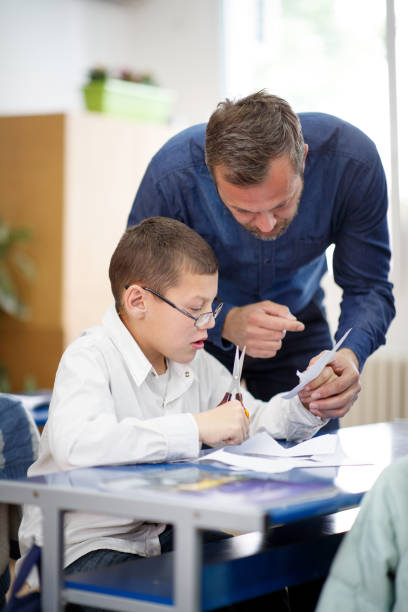Teacher helping special needs boy on his craft skills Male special education teacher working with a special needs elementary age boy, he is explaining him how to cut paper following pattern special education stock pictures, royalty-free photos & images
