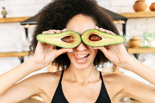 Attractive young multi ethnic woman with an afro hairstyle covered her eyes with halves of avocado. Funny picture with healthy food. Keto diet concept