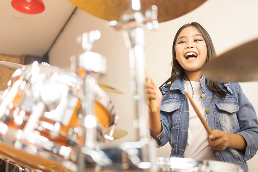 A cute Asian elementary school girl with long hair and a denim jacket laughing happily while playing a drum in a music classroom.