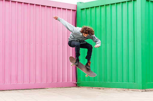 Young Man Jumping With Skateboard Against Corrugated Iron Wall full length by day.