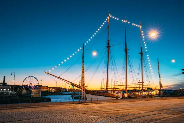 Photo of Helsinki, Finland. Old Wooden Sailing Vessel Ship Is Moored To The City Pier, Jetty. Unusual Cafe Restaurant In City Center In Lighting At Evening Or Night Illumination