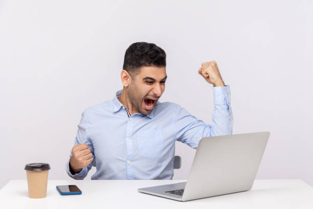 istock Successful delighted businessman sitting office workplace working on laptop, raising hands shouting for joy 1256493479