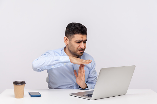 Need break! Upset male employee sitting office workplace, looking imploring at laptop screen and showing time out, asking pause in online conference. indoor studio shot isolated on white background