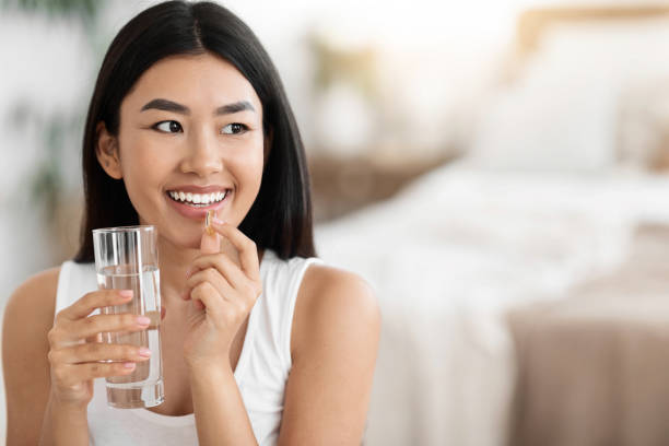 幸せなアジアの女性はピルを飲み、水のグラスを持っています - vitamin pill nutritional supplement capsule antioxidant ストックフォトと画像