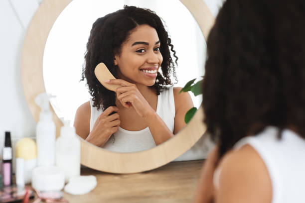 Attractive African Woman Combing Her Hair In Front Of Mirror At Home Attractive African Woman Combing Her Curly Hair In Front Of Mirror At Home, Smiling And Enjoying Her Beauty, Selective Focus Combing stock pictures, royalty-free photos & images