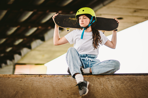 Teenage girl in helmet and stylish clothes posing on half pipe ramp an outdoor skate park. Beautiful kid female model skateboarder with skate board in urban extreme park. Schoolgirl after school.