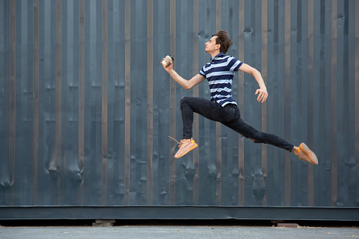 Jumping young buinessman in front of city building wall, on the run in jump high. Drinking coffee, moving to daily routine inspired and sportive. Young ballet dancer in casual clothes and sunshine.