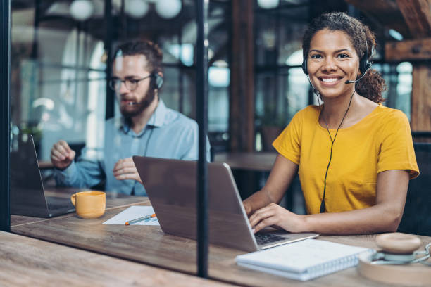 A smiling woman wearing a yellow shirt and a headset sits at a wooden table with a laptop, while a man in the background, wearing glasses and a blue shirt, is focused on his laptop, with a yellow coffee cup and stationery in front of her.