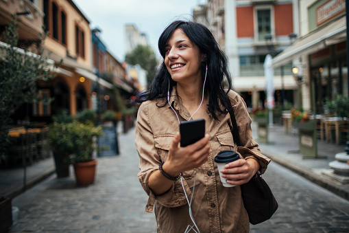 Smiling brunette holding her phone and a coffee while walking down a town street
