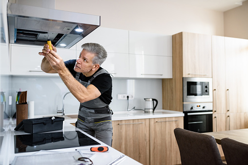 Aged repairman in uniform working, fixing broken kitchen extractor fan using screwdriver. Repair service concept. Selective focus. Horizontal shot