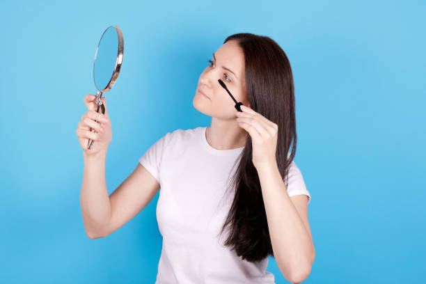 istock Girl looking at mirror and using maskara. Isolated on blue background. 1256533336