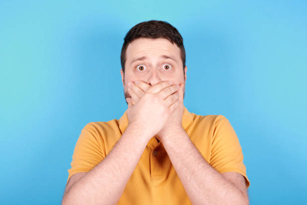 istock Brunette man scared, holds his hands ove mouth. Isolated on blue background. 1256533767