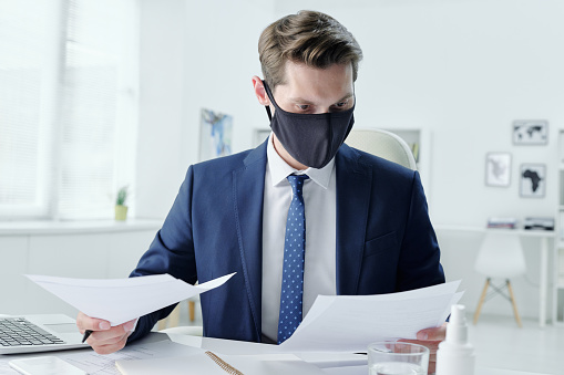 Modern young businessman in cloth mask sitting at table in office and reading contract while examining terms and conditions