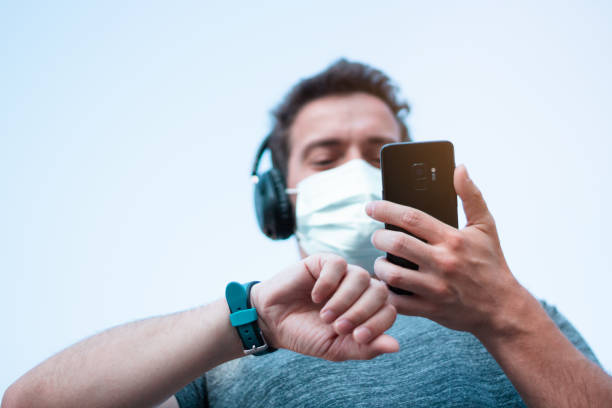Low angle shot of handsome man in a medical mask listening music and looking at his cell phone before he starts running. Coronavirus pandemic situation. stock photo