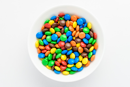 Group of small mixed colored chocolate coated candies in a white bowl on a table, isolated on white background, top view or flat lay photo of yellow, orange, green, blue, red and brown sweets