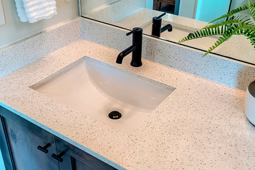 Bathroom white countertop with single basin undermount sink and black faucet. Cabinets, hanging towel, potted plant, and wall mirror can also be seen inside the room.