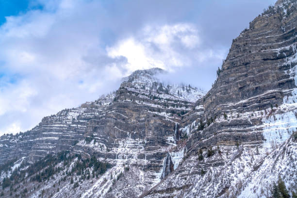 Cloudy blue sky over the scenic Provo Canyon with steep and rugged slopes. Snowy evergreens and frozen water can be seen on the mountain terrain in winter.