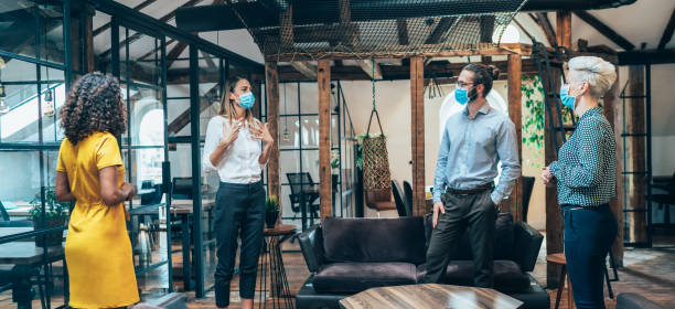 Place of work Colleagues in the office standing in distance and talking while wearing medical face mask during COVID-19 social distancing photos stock pictures, royalty-free photos & images
