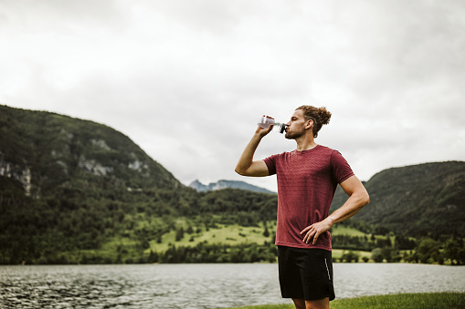 Young man in red t-shirt hydrating from water bottle. It's after sunset and there are lake and hills in the background. Copy space.