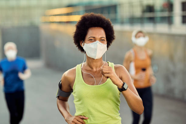 Jogging with a social distancing! Happy African American female runner wearing protective face mask while jogging outdoors during coronavirus epidemic. There are people in the background. illness prevention photos stock pictures, royalty-free photos & images