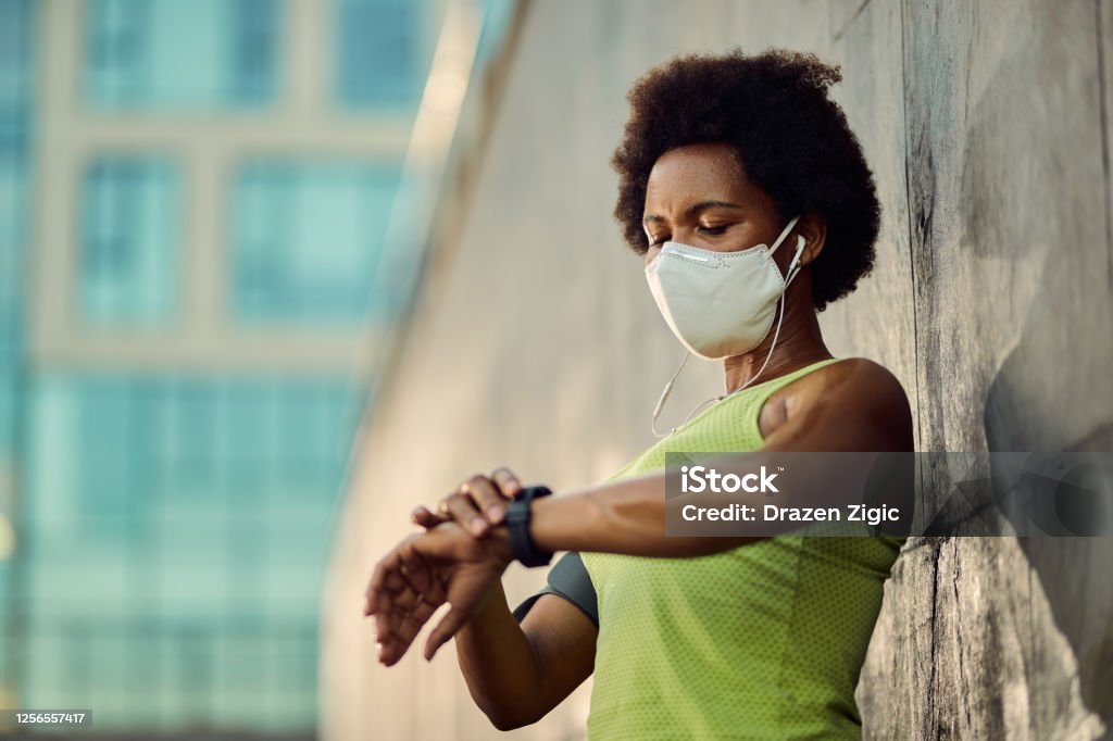 African American female runner with face mask using fitness tracker outdoors. Black athletic woman with protective face mask using smart watch while checking her heart rate after running outdoors. Running Stock Photo