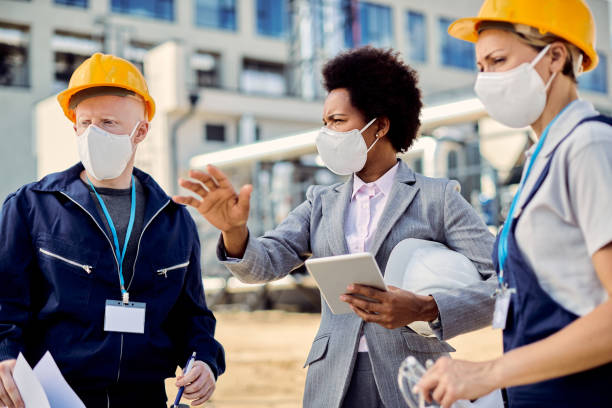 Black female architect and civil engineers with protective face masks talking at construction site.