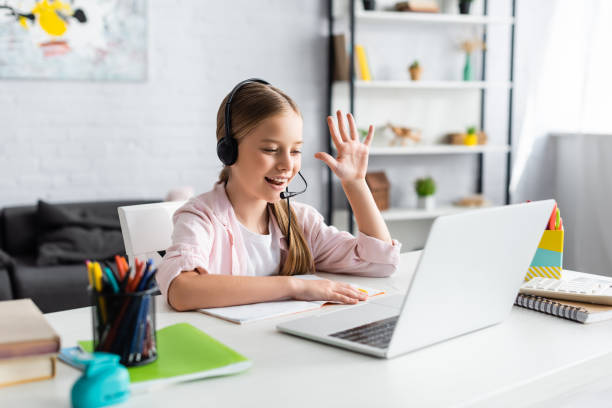 istock Selective focus of positive kid in headset waving hand during webinar at home 1256567257