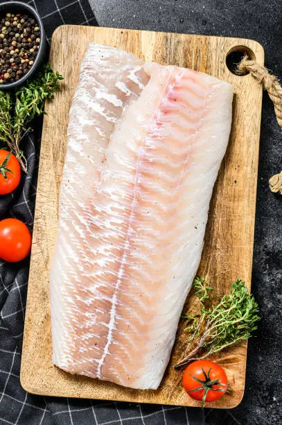 Photo of Raw cod fillet with and herbs on rustic wooden cutting board.  Black background. Top view