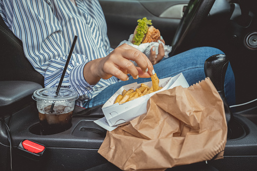 An unrecognizable woman is sitting in the driver's seat with her hands fully busy with the take out lunch. Tasty cheeseburger, cold soda, french fries... What else you could ask for...on a cheat day?