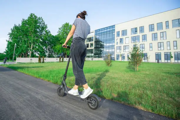 A rear view of an unrecognizable person riding on a scooter. She is spending a pleasant afternoon in black jeans, white sneakers and a bun in her hair.