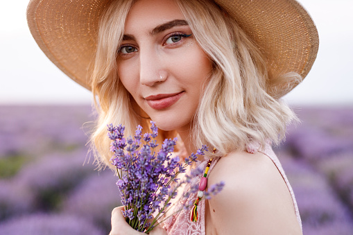 Side view of pretty fashionable gentle blonde female in straw hat standing in blooming field, with lavender flowers in hand and looking at camera with smile